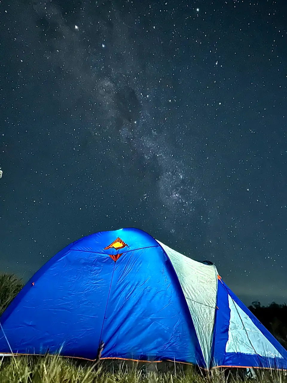 Barraca azul acesa sob o céu estrelado e a Via Láctea, no gramado do camping