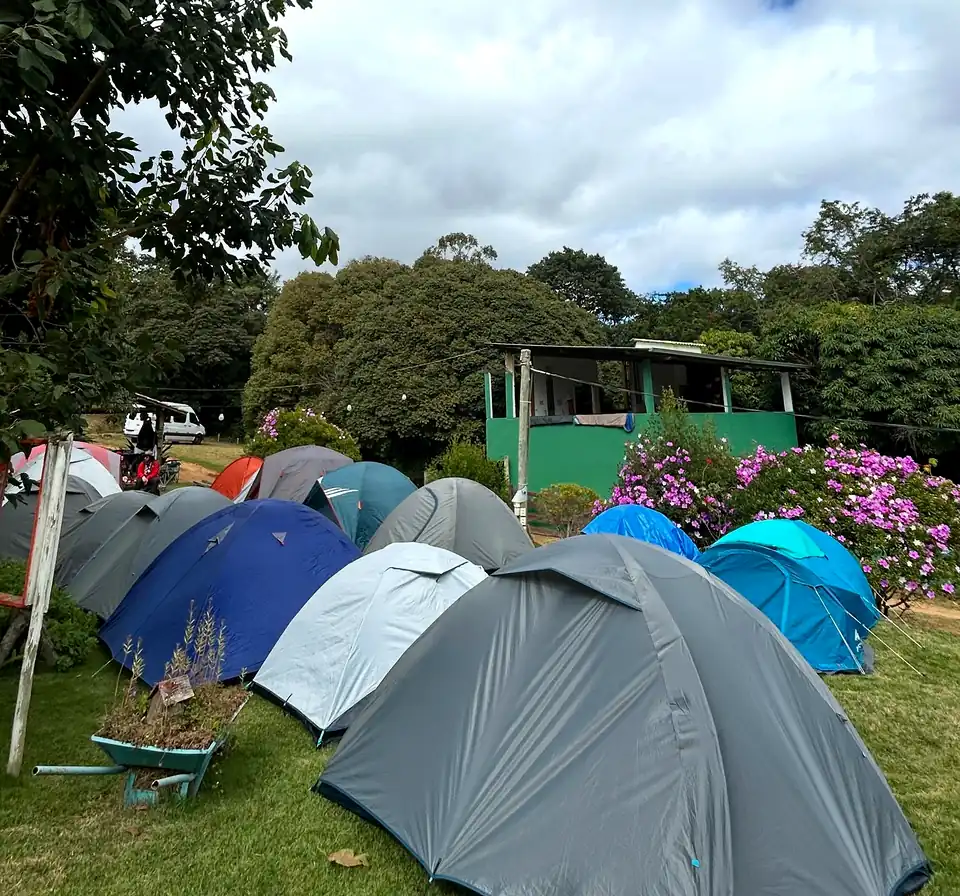 Barracas armadas no gramado do camping, com flores roxas e a casa verde ao fundo