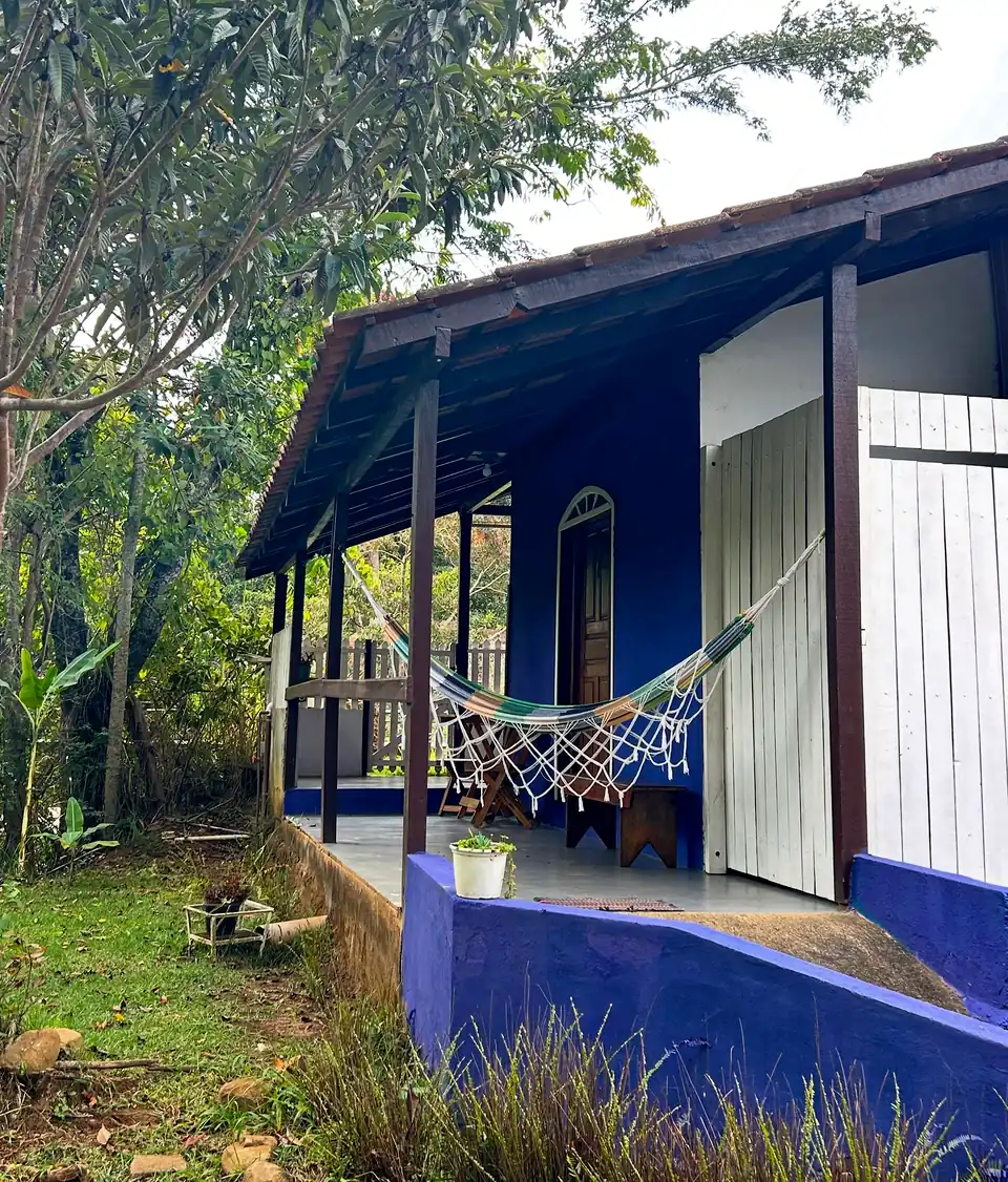 Chalé azul com varanda de madeira, rede branca e janelas de veneziana, cercado de árvores