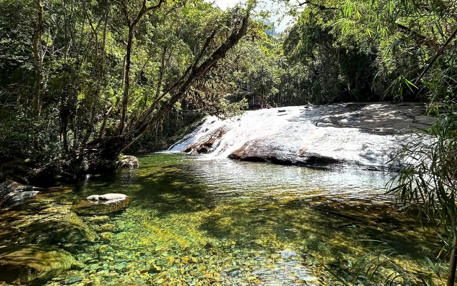 Poço de água verde e cristalina no rio, entre pedras e mata fechada, com a correnteza descendo pela pedra